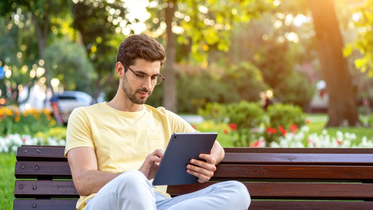 Young Man Using Digital Tablet on Wooden Bench in Sunny Park